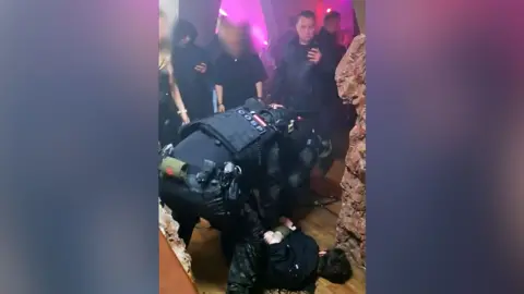 A member of Russian law enforcement is leaning over a person on the floor of the party, with their hands held behind their back, as other guests watch. The nightclub has pink neon lighting.