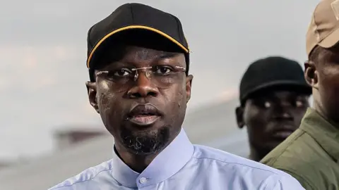 AFP via Getty Images A head and shoulders shot of Ousmane Sonko wearing glasses and in a black cap and buttoned-up blue shirt.