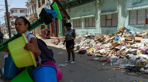 Magdalena Chodownik/Anadolu via Getty Images Two women stand in a street in Havana in which piles of rubbish have accumulated. There are empty cardboard boxes, bin bags and general refuse in a mountain at one side of the street. One of the women is carrying plastic buckets and brooms. Another stands in the middle of the street pointing at something. 