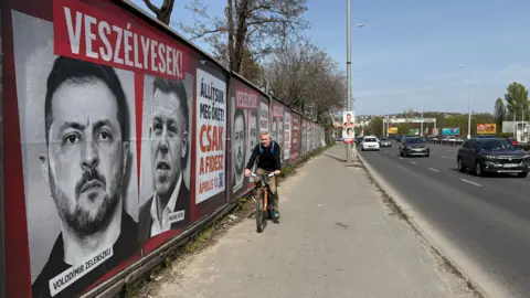 A man rides a bike past a wall along a highway in Hungary – plastered with noir headshots of Volodymyr Zelensky and Péter Magyar.