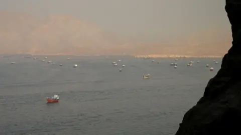 Reuters The Strait of Hormuz is shown from a dsitance w several tankers and freight boats. There are mountains in the foreground and in the distance.
