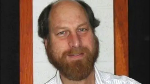 CalTech Carl Grillmair is seen smiling for a photo. He has a beard and is wearing a collared shirt 
