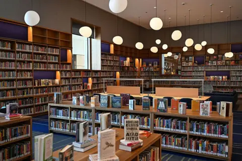 Joshua Lott for The Washington Post via Getty Images A room is lined with bookshelves and filled with book displayed made of light wood. There is a blue and green carpet and irregular orb lights hang from the ceiling.