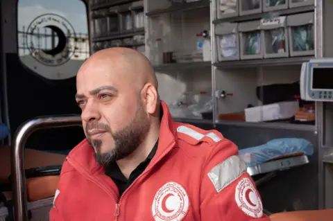 Paramedic Hassan Fouqha sits on the edge of his open ambulance bay door in a red medic jacket, looking straight ahead.