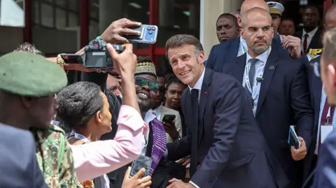 AFP via Getty Images Macron posing for a selfie as a man smiles and holds up his mobile phone. Others near them are holsing cameras and behind Macron is his security. Macron and his entourage are walking out of a building.