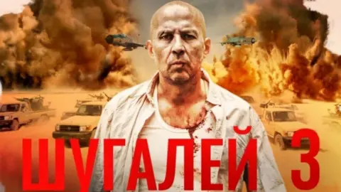 Youtube/ Aurum Production A man wearing a white shirt over a white tank top with traces of blood is standing in front of a scene of fighting in a dessert. 