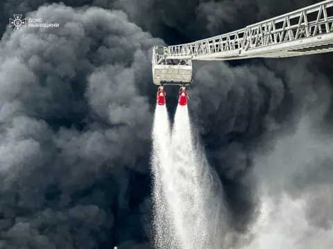 DSNS Thick dark grey smoke billows from the Brody oil hub as firefighters tackle an unseen fire with two hoses from an extended ladder.