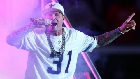 Getty Images Vanilla Ice sings into a microphone He wears a white NFL jersey with the number 31 on the front, a white baseball cap and a big silver neck chain.