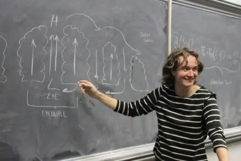 Courtesy of Aubrey Palmer Meteorology student Aubrey Palmer points to a weather diagram on a blackboard with a piece of chalk while smiling and facing a classroom. 