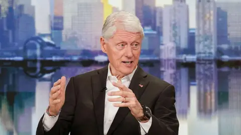 Getty Images Bill Clinton wears a black suit and a white shirt on a talk show tv set that has backdrop of New York City