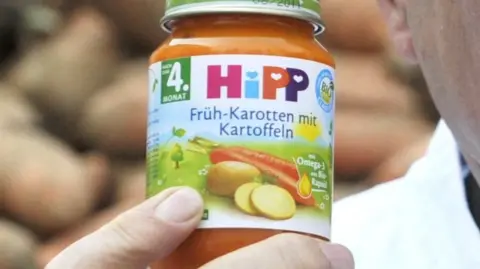 Getty Images A man holds a jar of HiPP carrot and potato baby food, with potatoes stacked in the background.