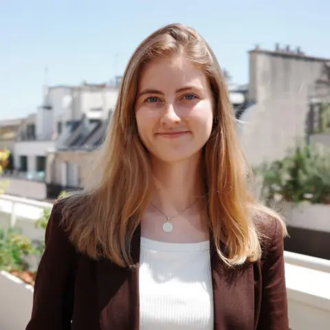 Emma Fourreau, stands on a sunny balcony with plants in the background. She is wearing a brown jacket which is open with a white ribbed top underneath. Emma Fourreau is smiling in the picture