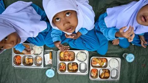 Getty Images Three Indonesian school children wearing blue uniforms and white veils, looking up, as they eat a free lunch on metal trays
