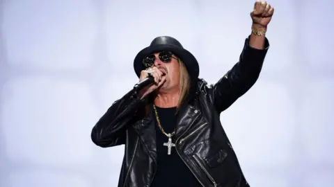 Getty Images Musician Kid Rock sings into a microphone and lifts a fist. He is wearing a black leather jacket, T-shirt, hat and sunglasses and a large silver cross on a chain.