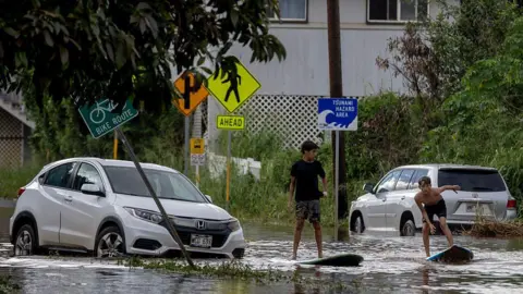 San Francisco Chronicle via Getty Images Two teenagers on surf boards in shallow flood waters on a street next to a disabled grey car. 