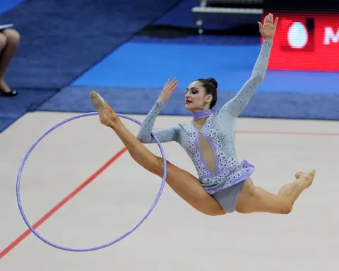 AFP via Getty Images Zeynab Javadli at a gymnastics competition in 2011. She is wearing a blue and purple leotard embellished with jewels. The photo captures her jumping in the air doing a split kick with her front leg and arm going through a large hoop.