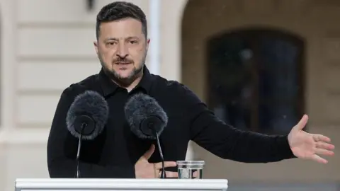 EPA Volodymyr Zelensky stands at a white podium in front of a building. The podium has the Ukrainian flag on it and two microphones and the president is dressed in black and gesturing to the right side of the image with his hand