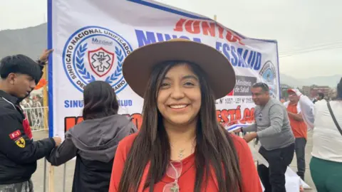 Janeth wearing a red dress and top, with long hair seen under a hat. She is standing in front of a campaign poster.
