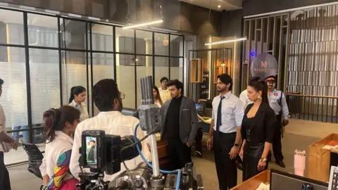 Don Vanzara Showbiz LLP A group of actors wearing office formals shoot a scene on a set which resembles a modern office space. There is a person wearing a security guards uniform standing at the back as cameras placed vertically record the scene. 