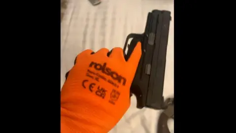 Counter Terrorism Policing An orange-gloved hand holding a black pistol. An old revolver is lying on the bed behind. 