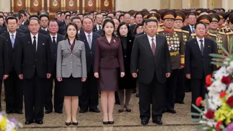 Reuters At a tribute at Pyongyang mausoleum the Kumsusan Palace of the Sun, in front of several rows of besuited and uniformed dignitaries, some with many medals on their chests, Ri Sol Ju, in a buttoned-up grey jacket, black skirt and shoes, is next to Kim Ju Ae, in matching shoes, who is next to Kim Jong Un, in a buttoned-up dark grey suit and white shirt, all standing to attention. Ju Ae wears a burgundy suit that matches her mother’s scarf and father’s tie. Colourful bouquets are in the foreground.