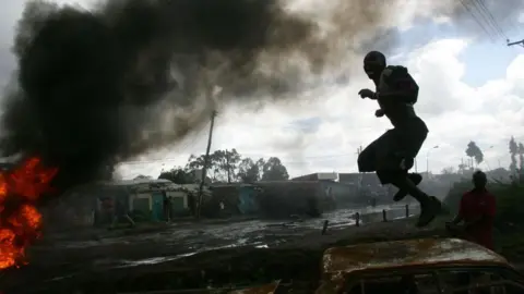 AFP via Getty Images A silhouetted figure jumps on the roof of a burnt vehicle on the right of the picture. Flames can be seen on the left edge of the shot with thick, black smoke emerging from them. Market stalls can be seen the background. 