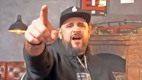 YouTube El Temach wears a black hoodie, a black and white cap and has a beard - he is pointing and shouting at the camera 