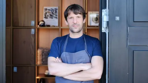 Getty Images Rene Redzepi, chef and co-owner of Danish restaurant Noma is pictured on May 31, 2021 in Copenhagen wearing a blue t-shirt and apron with his arms crossed.