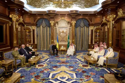 Reuters Ukraine's President Volodymyr Zelenskiy is black and the Crown Prince of Saudi Arabia, Mohammed bin Salman Al Saud is next to him. Advisors sit on either side in a grand meeting room with blue carpet