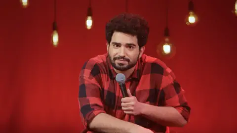 Samay Raina via YouTube Dressed in a black-red check shirt, Samay Raina looks into the camera while holding a mic in his left hand