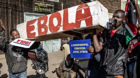 AFP via Getty Images Protesters carry a white coffin, with the word "Ebola" in red paint on the side. They are also holding placards saying - in Swahili - "we reject Ebola" and - in English: "Public trust requires transparency".