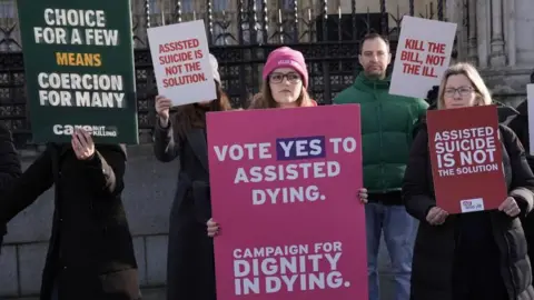PA Media An image showing protestors both for and against assisted dying in the UK. One holds a sign that says 'Vote YES to assisted dying'. Around her are several people holding signs that say 'Kill the bill, not the ill' and 'Assisted sucidie is not the solution'. 