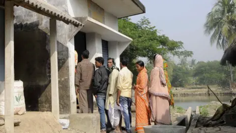 Hindustan Times via Getty Images Six people - four men and two women - standing in queue outside a public sector bank's branch in Sunderban, India. The picture is from 2016. 