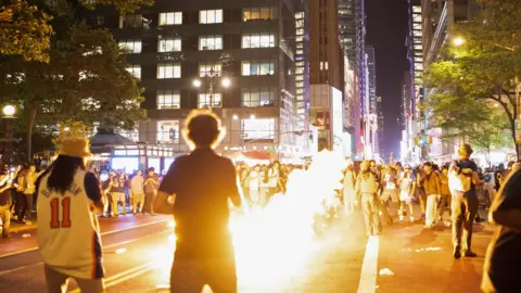 REUTERS/Christian Monterrosa A man walks away from the camera toward a browd on a New York street - there is fire in front of him 