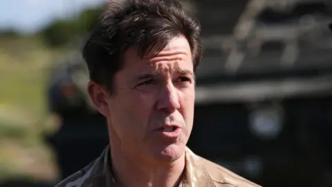 PA Media Major General Tom Bewick, who is middle-aged and has short brown hair, is seen squinting in the sun as he speaks to journalists at RAF Akrotiri 