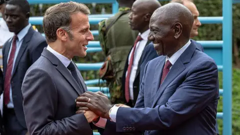 AFP via Getty Images Macron and Ruto are facing each other, smiling and clasping hands. They are outdoors and there are four men behind them