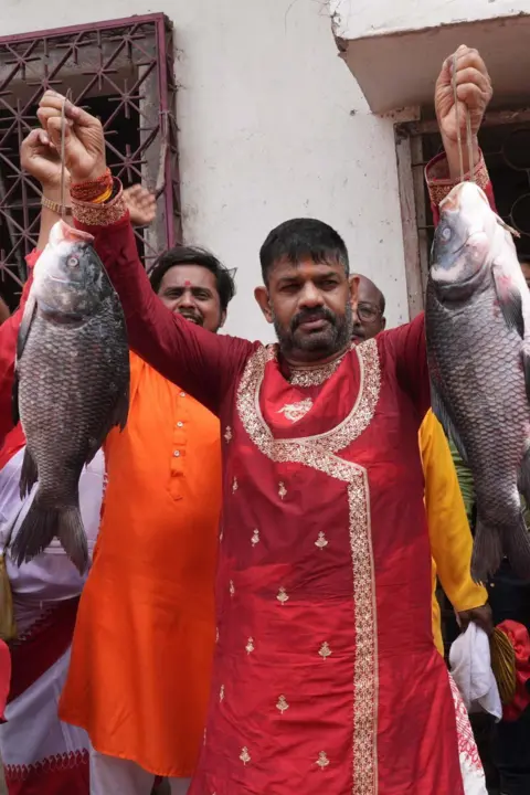 A man wearing a red kurta holds up a fish each in either hand while looking at the camera