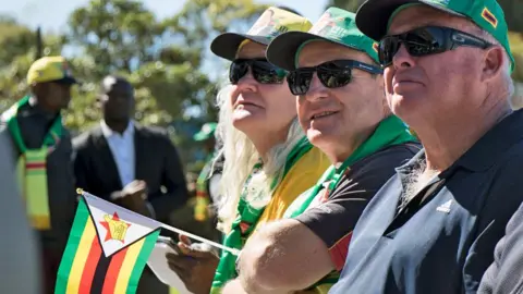 AFP via Getty Images Two men and one woman in green hats and one holding a Zimbabwean flag attend a political campaign meeting held by President Emmerson Mnangagwa in July 2018 for members of the white farming and business community.