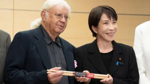 EPA Japanese Prime Minister Sanae Takaichi poses with Ian Paice. They hold a set of signed drumsticks between them. Takaichi has short black hair and wears a black blazer over a black top. Paice has long white hair, tied back, and wears glasses, a black blazer and a grey buttoned shirt.
