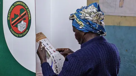 AFP via Getty Images A person stands inside a polling booth in a classroom-like setting, marking a ballot. The room has worn walls, wooden desks, and hand‑drawn posters displayed on the walls. 