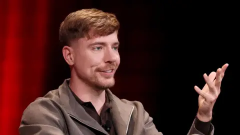 Getty Images MrBeast, real name Jimmy Donaldson, at an event. He is wearing a brown t-shirt and zip top, with his left arm gesturing as he speaks.