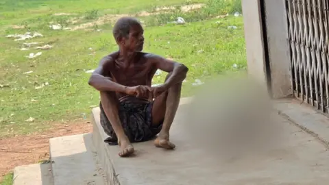 BBC Hindi A man wearing a dark printed dhoti - which is a cloth tied around the waist and covering until the knee - sits on stairs. With him are the remains of his sister - which have been blurred. There is green grass in the background and a metal gate on one side. 