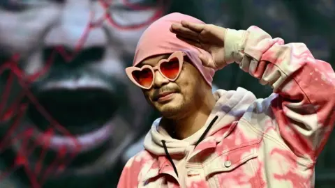Getty Images Namewee wearing a pink beanie, pink heart-shaped sunglasses and a pink denim jacket, raising his hand to his forehead in a saluting pose.