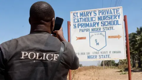 EPA/Shutterstock A policeman with his back to camera taking a photo with his phone of a sign pointing to St Mary's School in Papiri, Niger state - November 2025