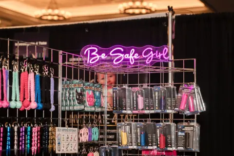 CrimeCon and RenownedPhotos A neon sign says "Be Safe Girl" and advertises rainbow-hued self-defense products like rape whistles