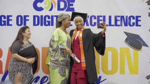 A graduate in black cap and gown holding a diploma takes a selfie with Fatima Bio in traditional African dress at a graduation ceremony, while a third woman smiles nearby