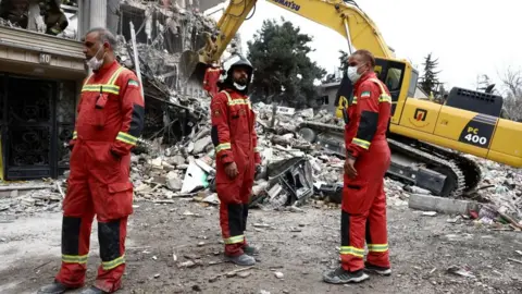 Reuters Three emergency workers in red uniforms stand near a yellow digger as it removes rubble