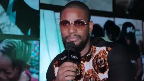 Alexx Ekubo via YouTube He is wearing a white tshirt with short black sleeves. The front has the repeated pattern of a roaring lion. He is wearing tinted sunglasses and is indoors. He is holding a mic.