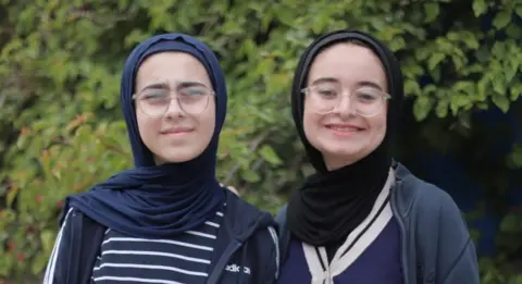 The Earth Prize Tala and Farah Mousa, two teeanged girls. Both have their heads covered in hijabs, wear glasses and navy zip-up jackets, and are smiling at the camera in front of a large green tree.