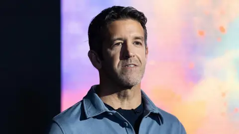 Getty Images John Ternus wears a blue shirt over a black t-shirt and stands in front of a colourful background of blue, purple, yellow, orange and pink 